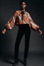 Load image into Gallery viewer, Model wearing a patterned blouse and black pants against a dark background