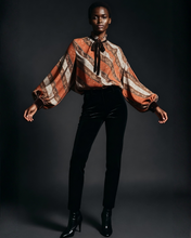 Load image into Gallery viewer, Model wearing a patterned blouse and black pants against a dark background
